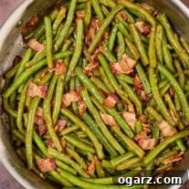 A sauté pan filled with cooked Southern style green beans with bacon, next to a wooden spoon and several scattered fresh beans.