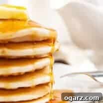 A side view of a stack of fluffy pancakes on a white plate, topped with a thick pat of butter and a drizzle of maple syrup.