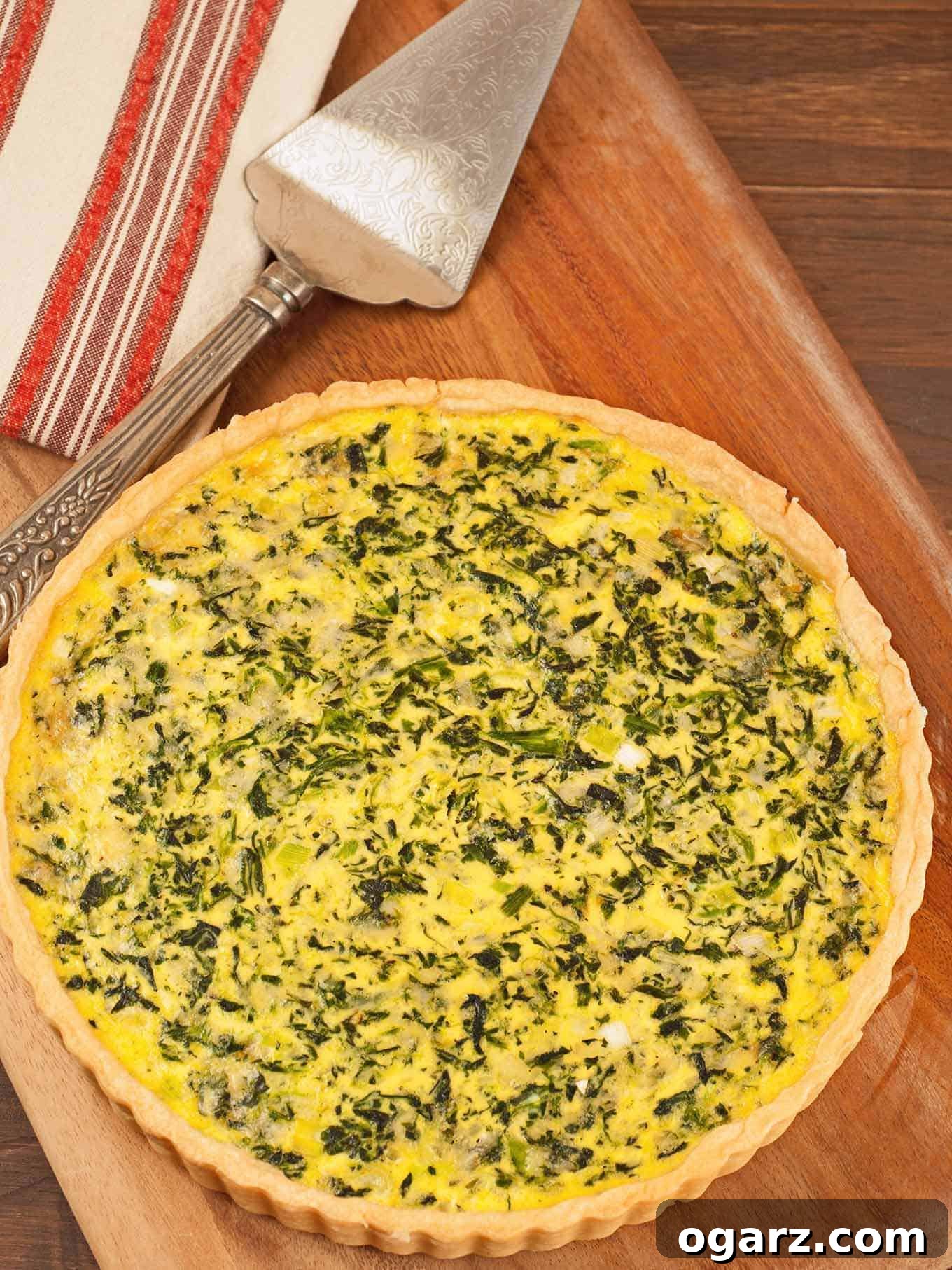 An overhead view of an uncut spinach and cheese quiche on a cutting board, a pie server and tea towel in the background.