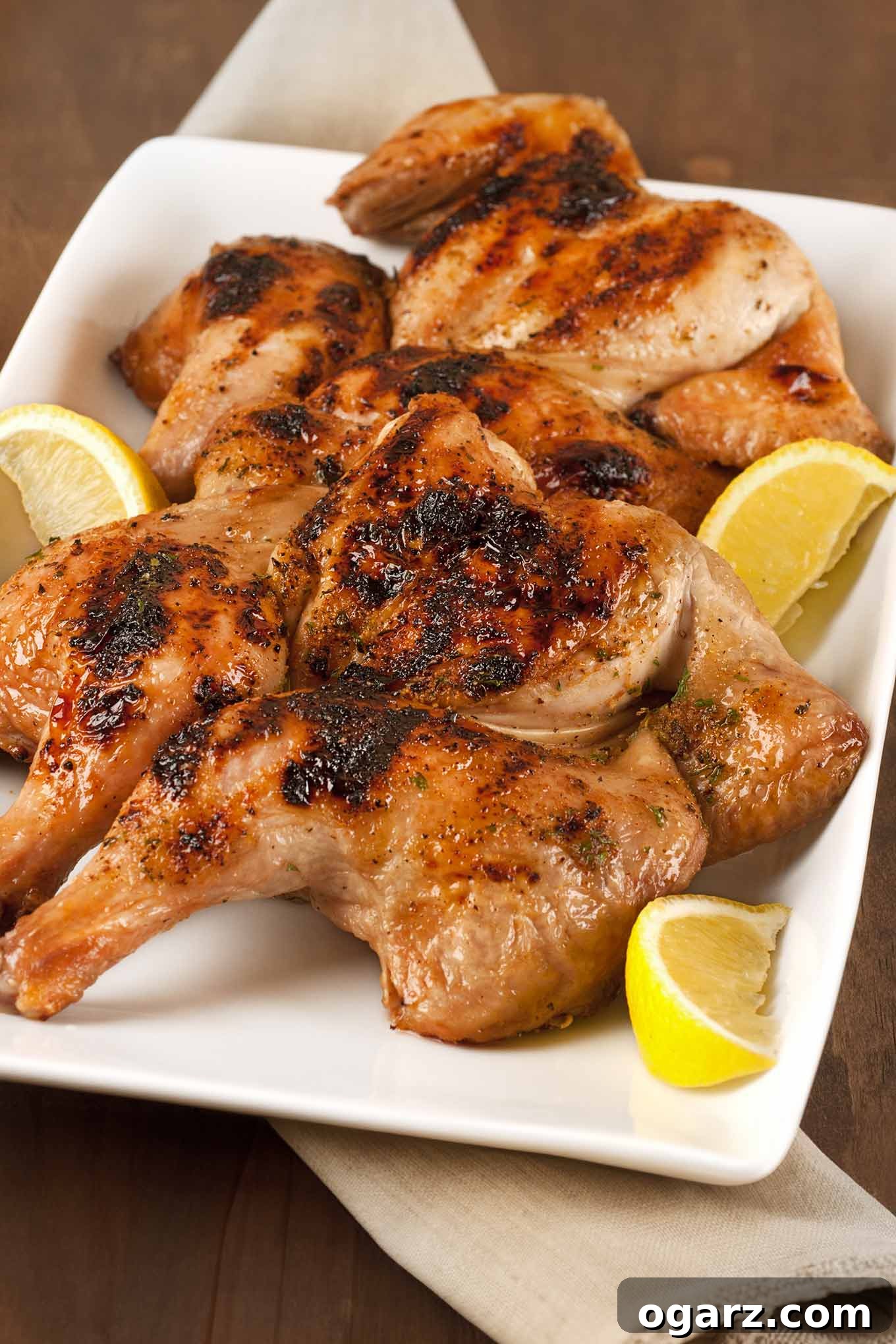 Golden Grilled Game Hens 2 Grilled spatchcocked, honey-glazed Cornish hens with charred skin on a white platter with lemon wedges on the side.