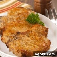 An oval plate with three breaded, oven-fried pork chops, garnished with parsley, a pepper mill and serving fork in the background.