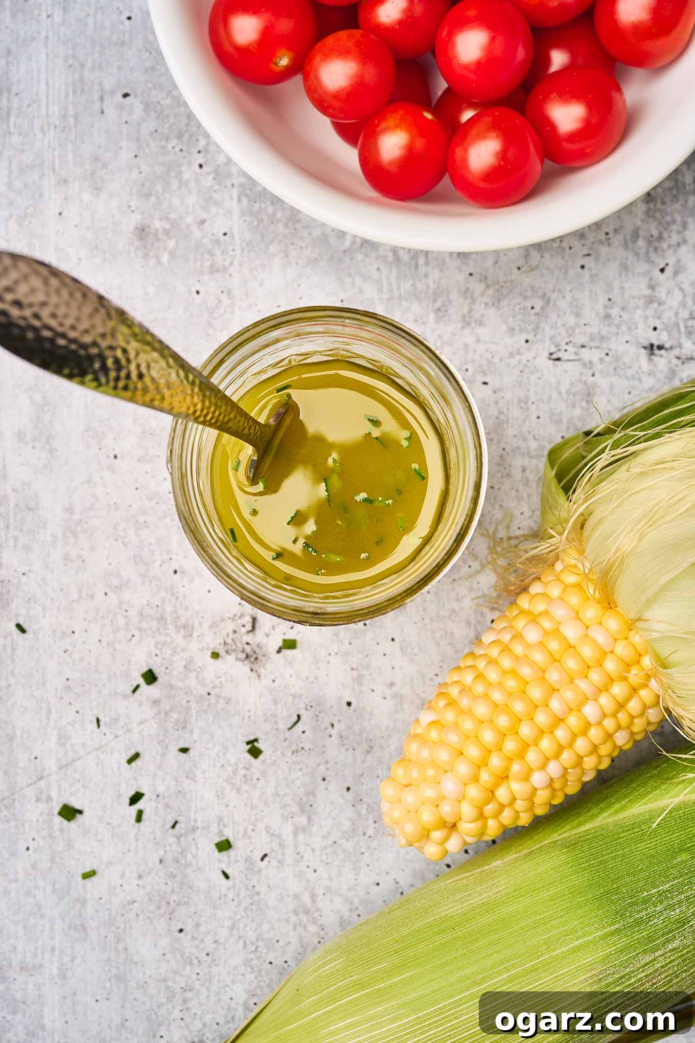 Sunshine Corn and Tomato Medley 4 Fresh corn on the cob in the husks, cherry tomatoes, and a jar of lemon, shallot, and chive vinaigrette on a gray cement background.