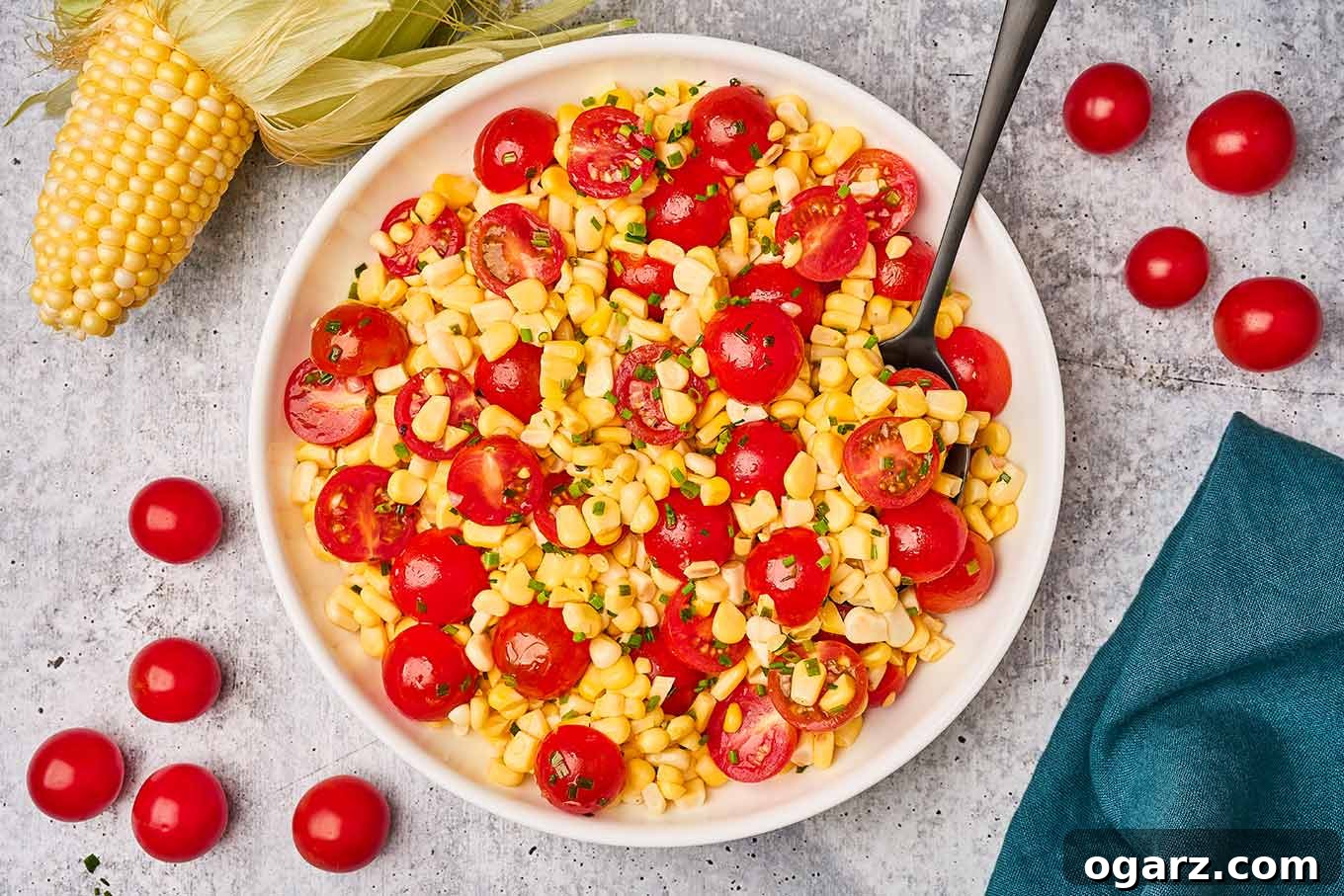 Sunshine Corn and Tomato Medley 5 An overhead view of corn and tomato salad in a serving dish, surrounded by extra salad ingredients on a gray cement background.