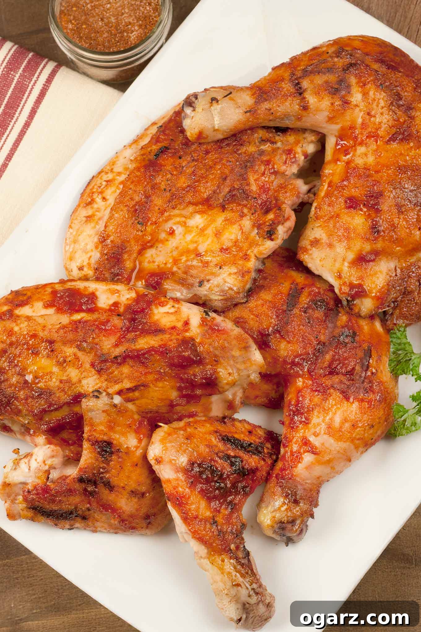 Slow Grilled Succulent Barbecue Chicken 2 A beautifully presented white serving platter laden with perfectly grilled BBQ chicken parts: succulent leg quarters, juicy breasts, and tender wings. A fresh sprig of parsley garnishes the side, and a jar of homemade BBQ spice rub stands invitingly in the soft-focus background, suggesting the flavorful journey ahead.