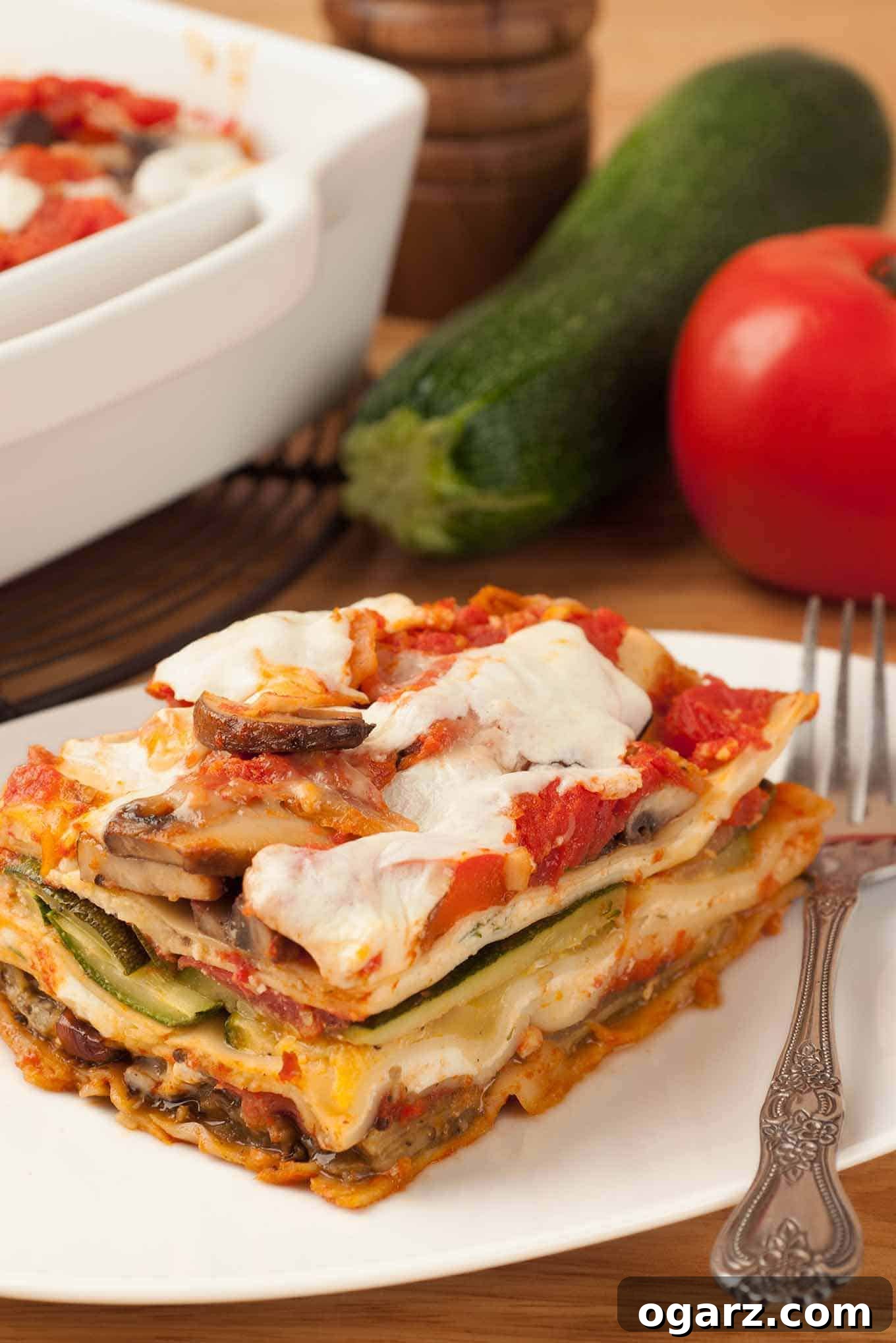 Garden Lasagna 2 A vibrant serving of homemade vegetable lasagna, featuring layers of pasta, rich tomato sauce, roasted zucchini, eggplant, mushrooms, and melted mozzarella cheese, presented on a white plate with a fork. In the background, fresh zucchini and tomatoes, alongside a full casserole dish of lasagna, suggest the bounty of fresh ingredients and the comforting nature of the meal.