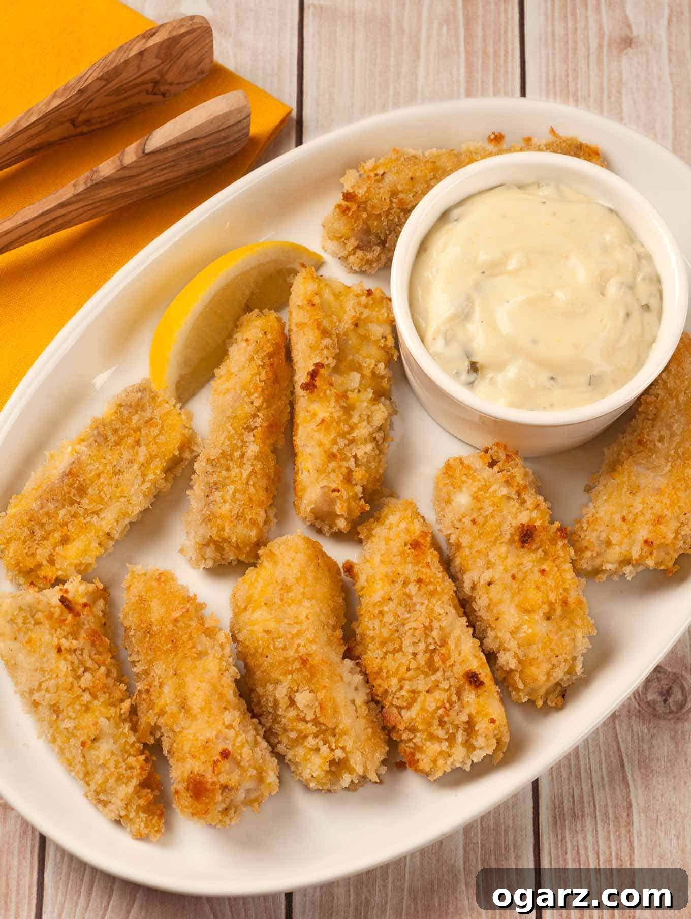 A serving platter of oven-fried fish sticks with a golden crispy coating, served with tartar sauce and lemon wedge. The perfect healthy alternative to store-bought frozen fish sticks.