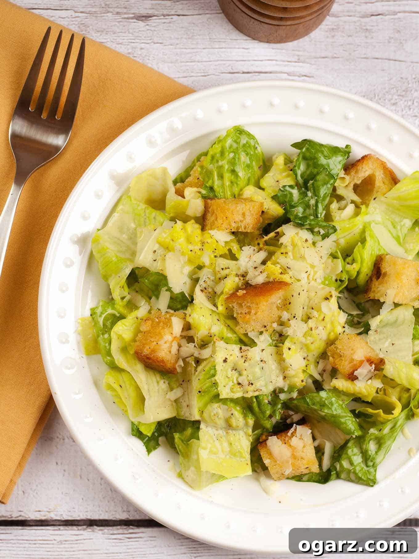 An overhead view of classic Caesar salad with crisp romaine lettuce, golden garlic croutons, freshly grated Parmesan cheese, and a generous sprinkle of black pepper on a pristine white plate, with a napkin and fork thoughtfully placed to the side, ready to be enjoyed.
