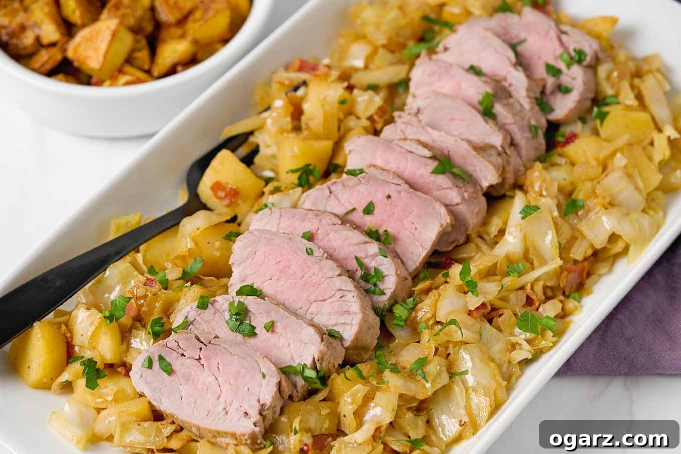 Apple and Cabbage Pork Tenderloin Braise 5 A beautifully composed white serving platter showcasing cider-braised pork tenderloin, artfully arranged on a bed of tender cabbage and sweet apples. A rich plum-colored napkin and a dish of golden roasted potatoes are elegantly placed to the side, completing the inviting meal.