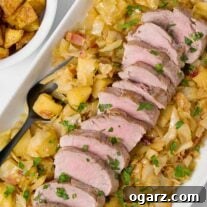 A platter of braised pork tenderloin, sliced and served on a bed of cabbage, apples, and bacon, with a bowl of roasted potatoes on the side.