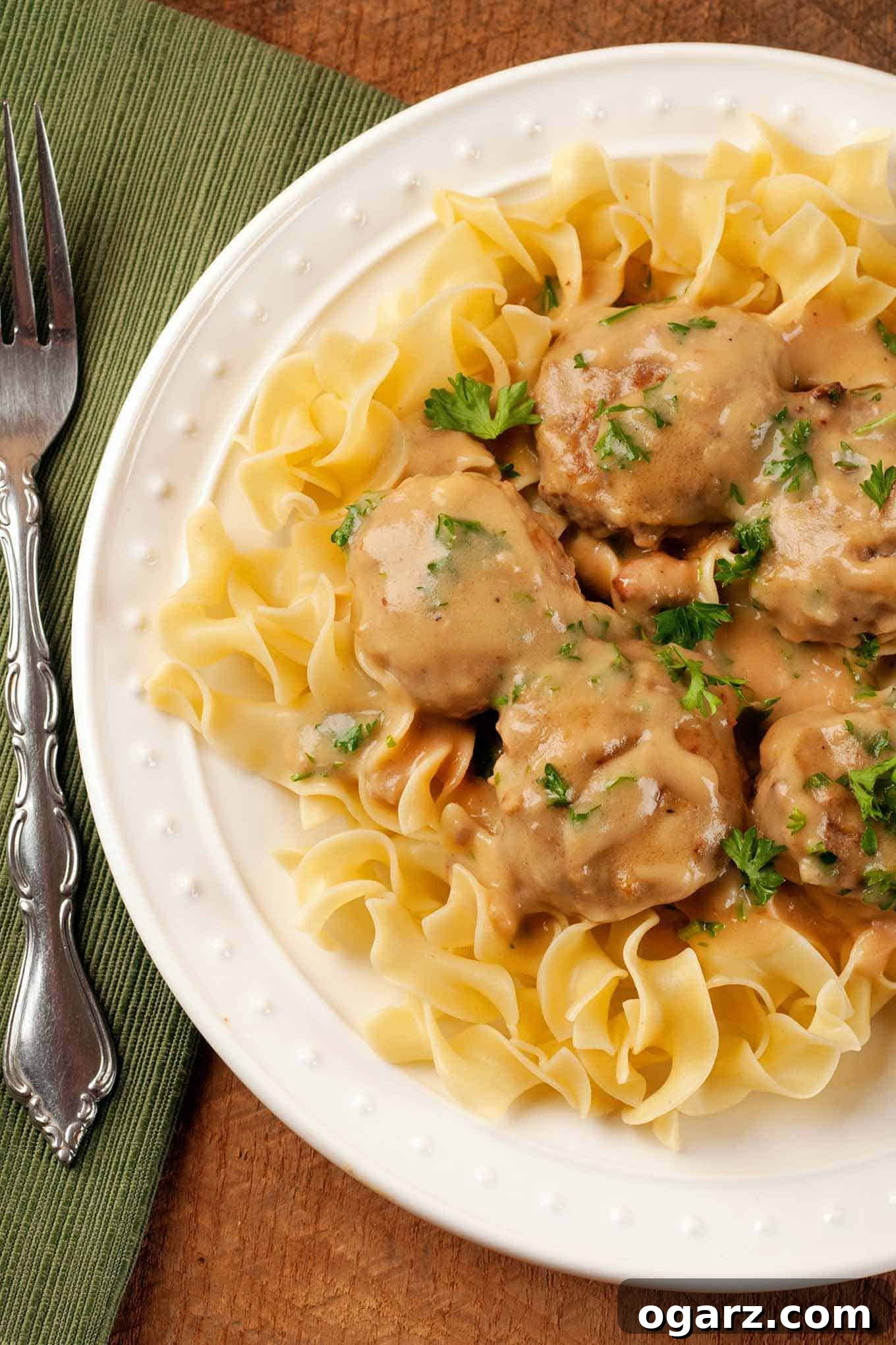 Homestyle Swedish Meatballs 2 An overhead view of a plate of classic Swedish meatballs served over tender egg noodles and garnished with fresh chopped parsley, with a napkin and fork neatly placed to the left.