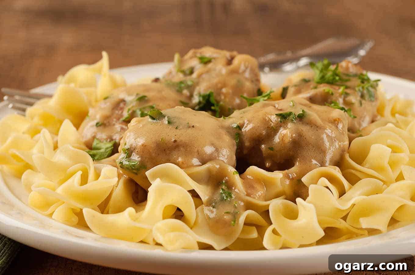 Homestyle Swedish Meatballs 3 A close-up view of succulent Swedish meatballs generously topped with vibrant chopped parsley, resting on a bed of glistening egg noodles, showcasing the luxurious creaminess of the accompanying sauce.