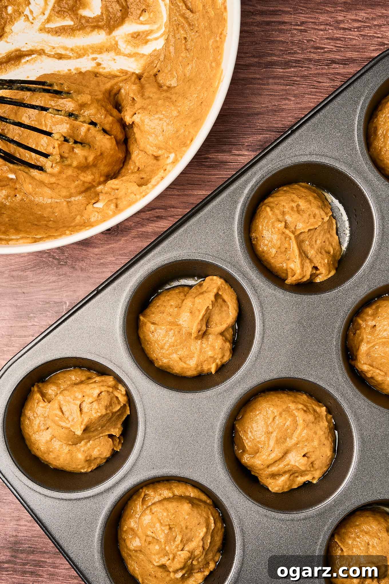 Pumpkin spice muffin batter spooned into a muffin tin and ready for baking, a bowl with more batter and whisk in the background.