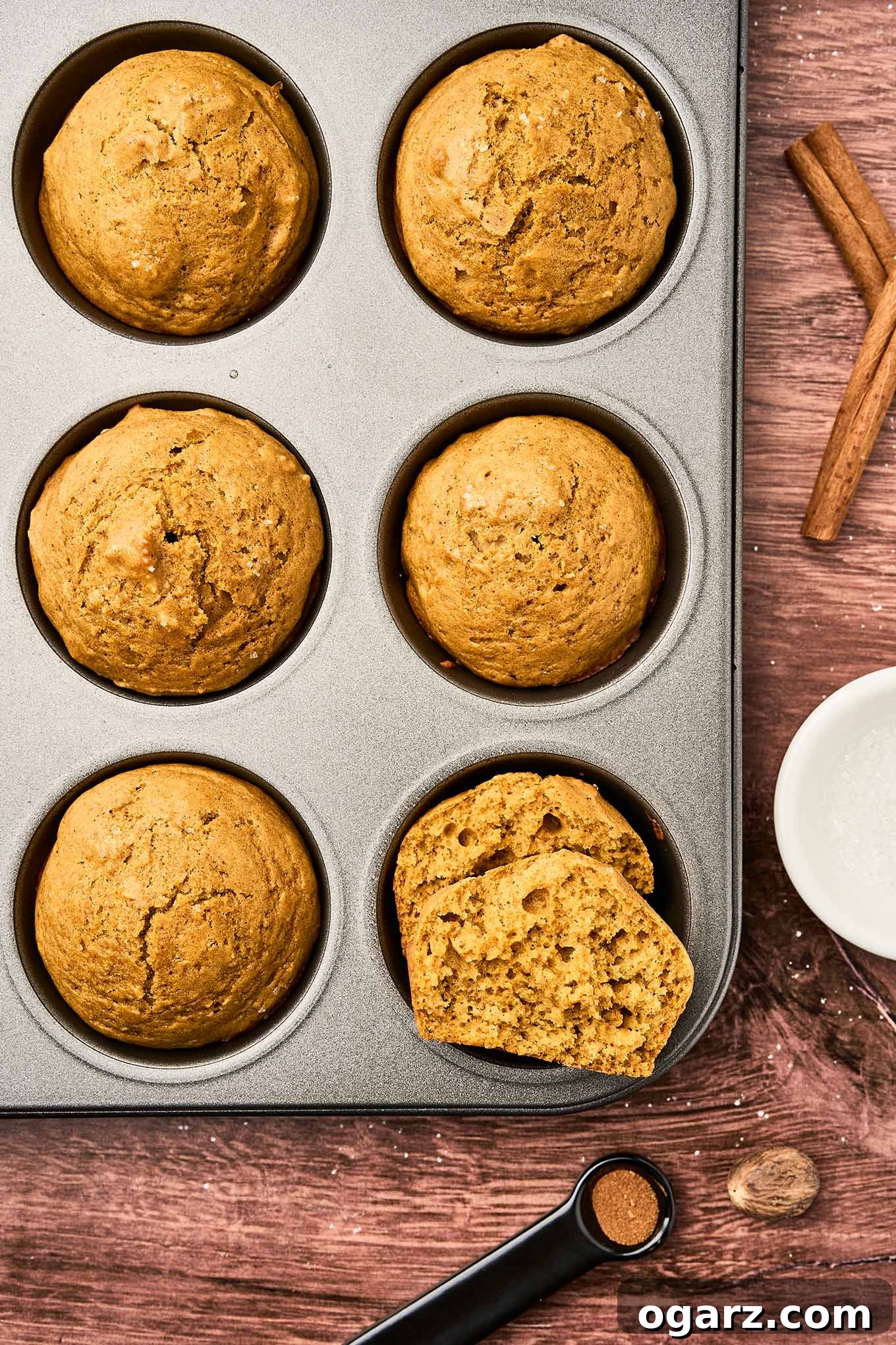 Golden brown pumpkin spice muffins cooling in a muffin tin, surrounded by cinnamon sticks, whole nutmeg, and a spoon of ground cloves, showcasing their rich aroma.