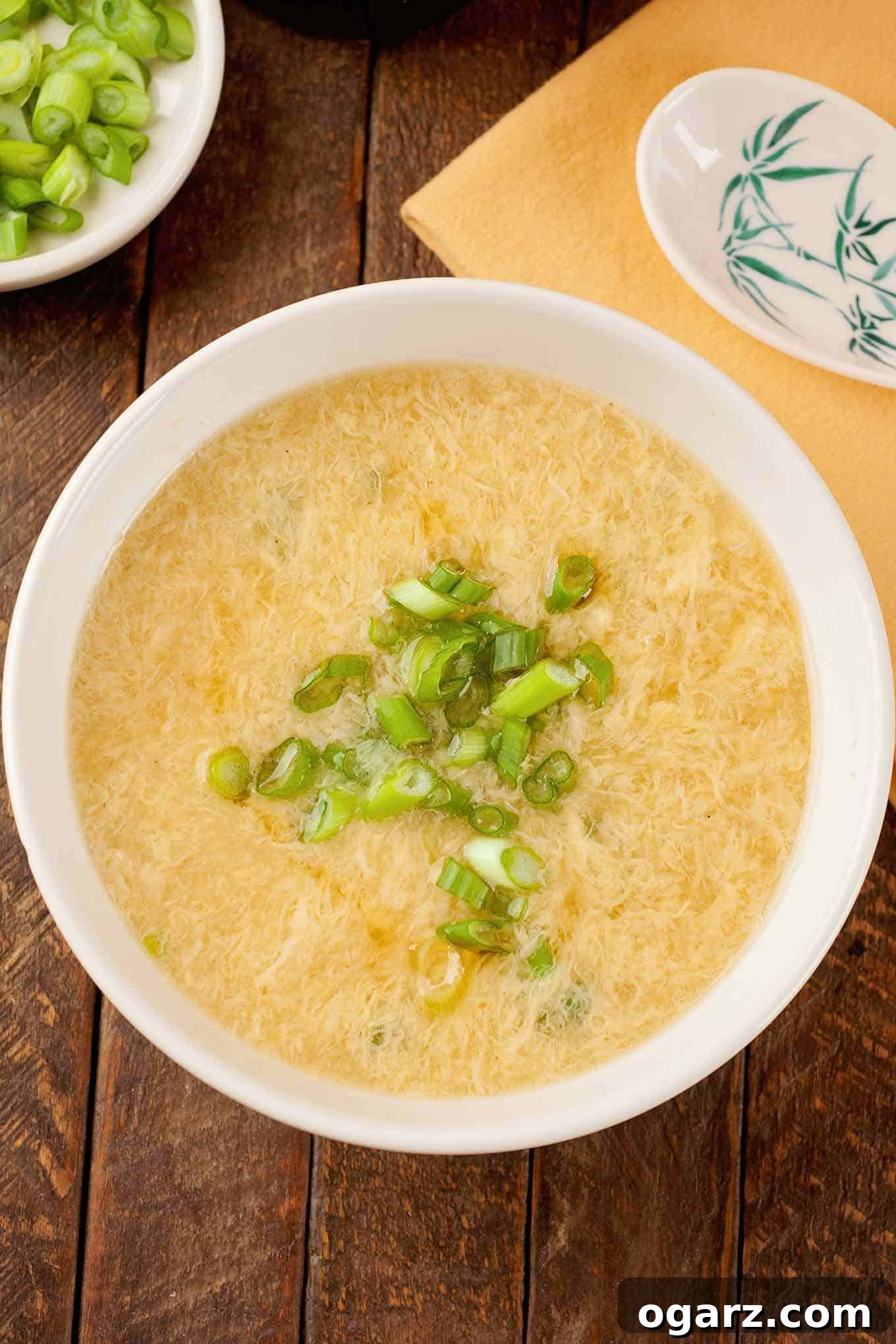 An overhead view of a bowl of egg drop soup garnished with scallions, a dish with more scallions, a napkin, and a ceramic spoon in the background.