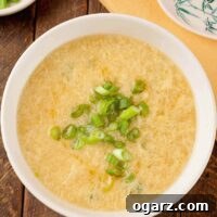 An overhead view of a bowl of egg drop soup garnished with scallions, a dish with more scallions, a napkin, and a ceramic spoon in the background.