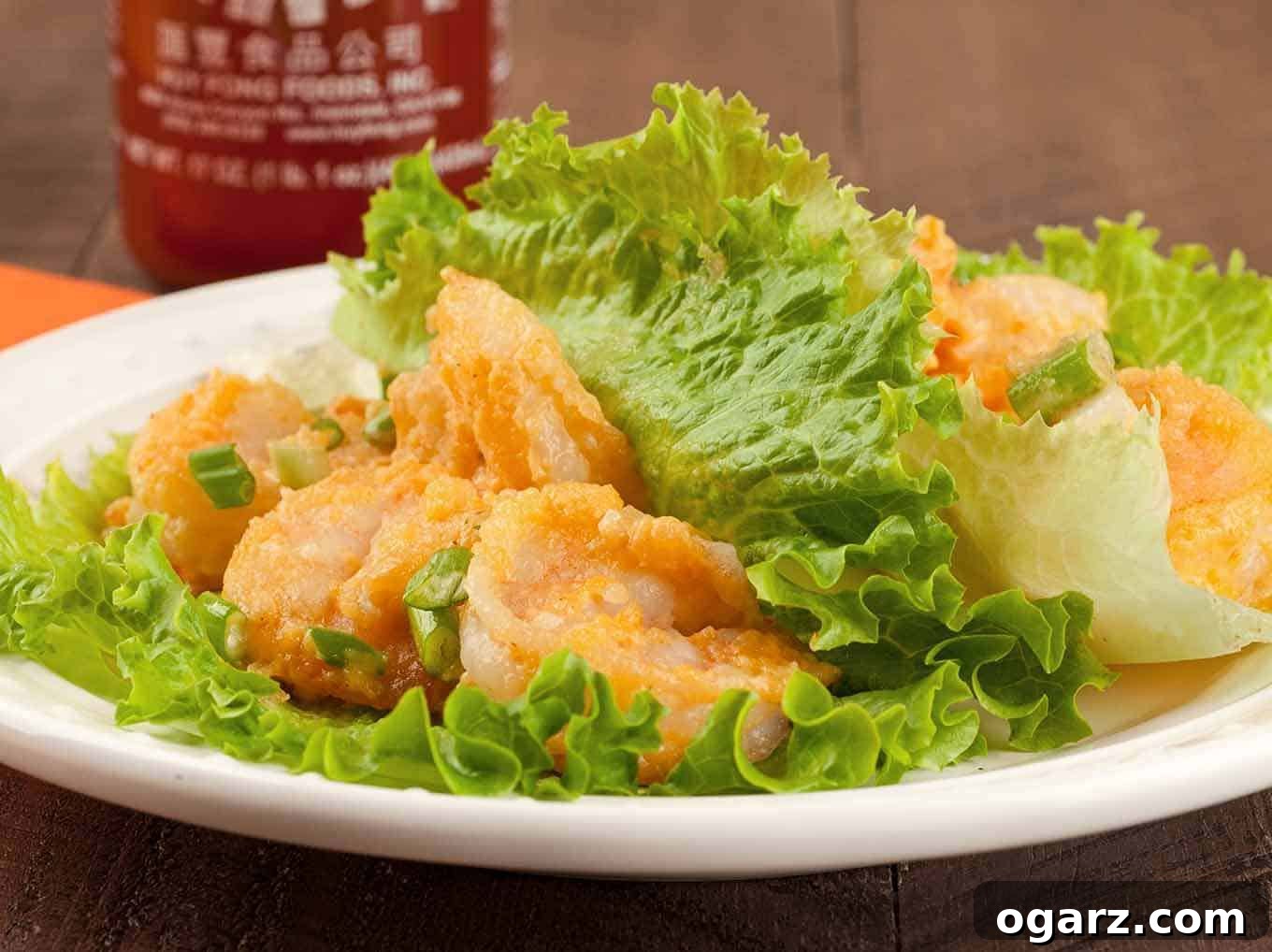 A close view of Bang Bang shrimp lettuce wraps on a white plate with a bottle of sriracha in the background, showcasing the vibrant sauce and fresh lettuce.