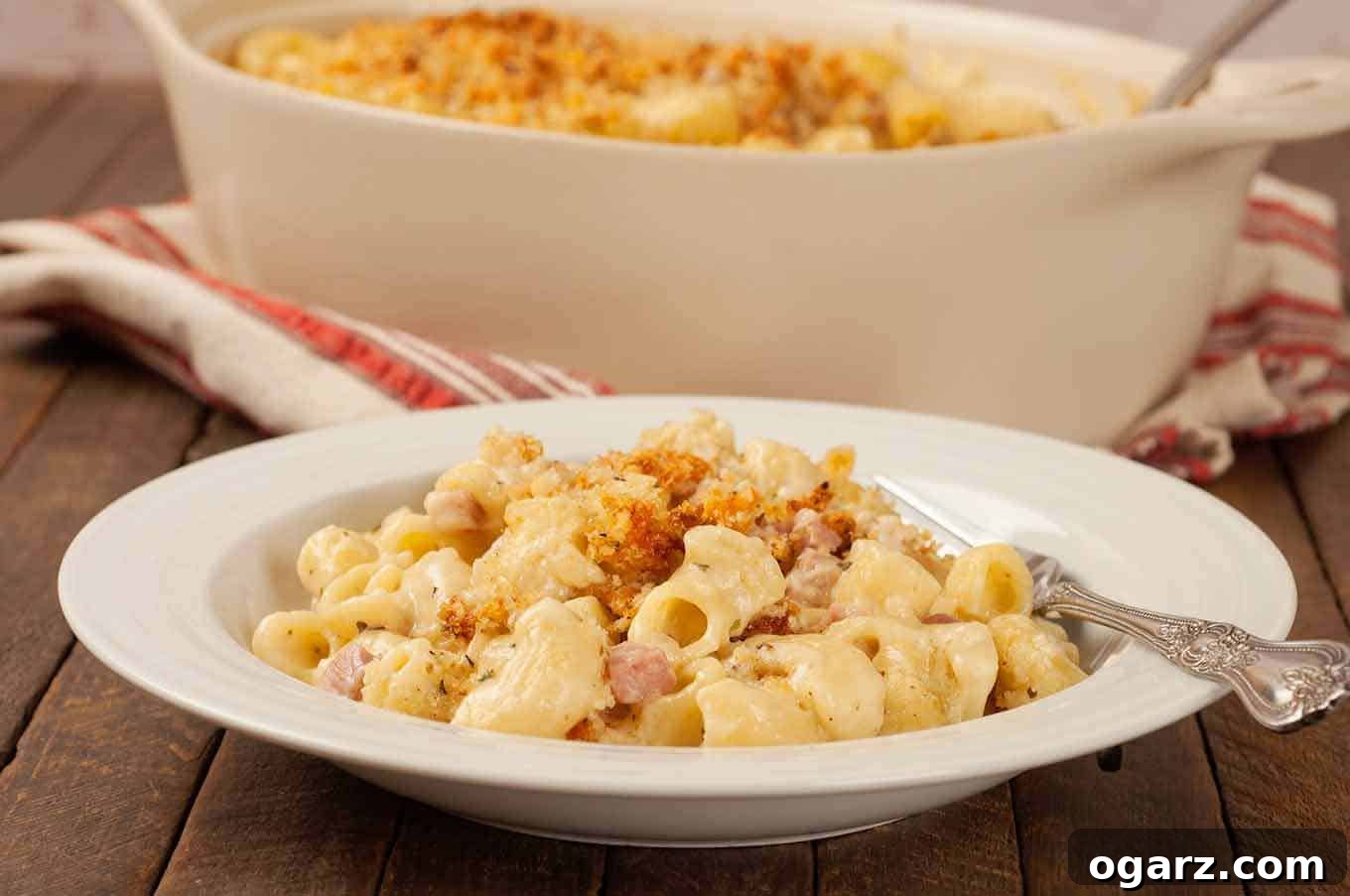 A shallow bowl filled with a serving of creamy macaroni and cheese, fork on the side and the whole casserole in the background.