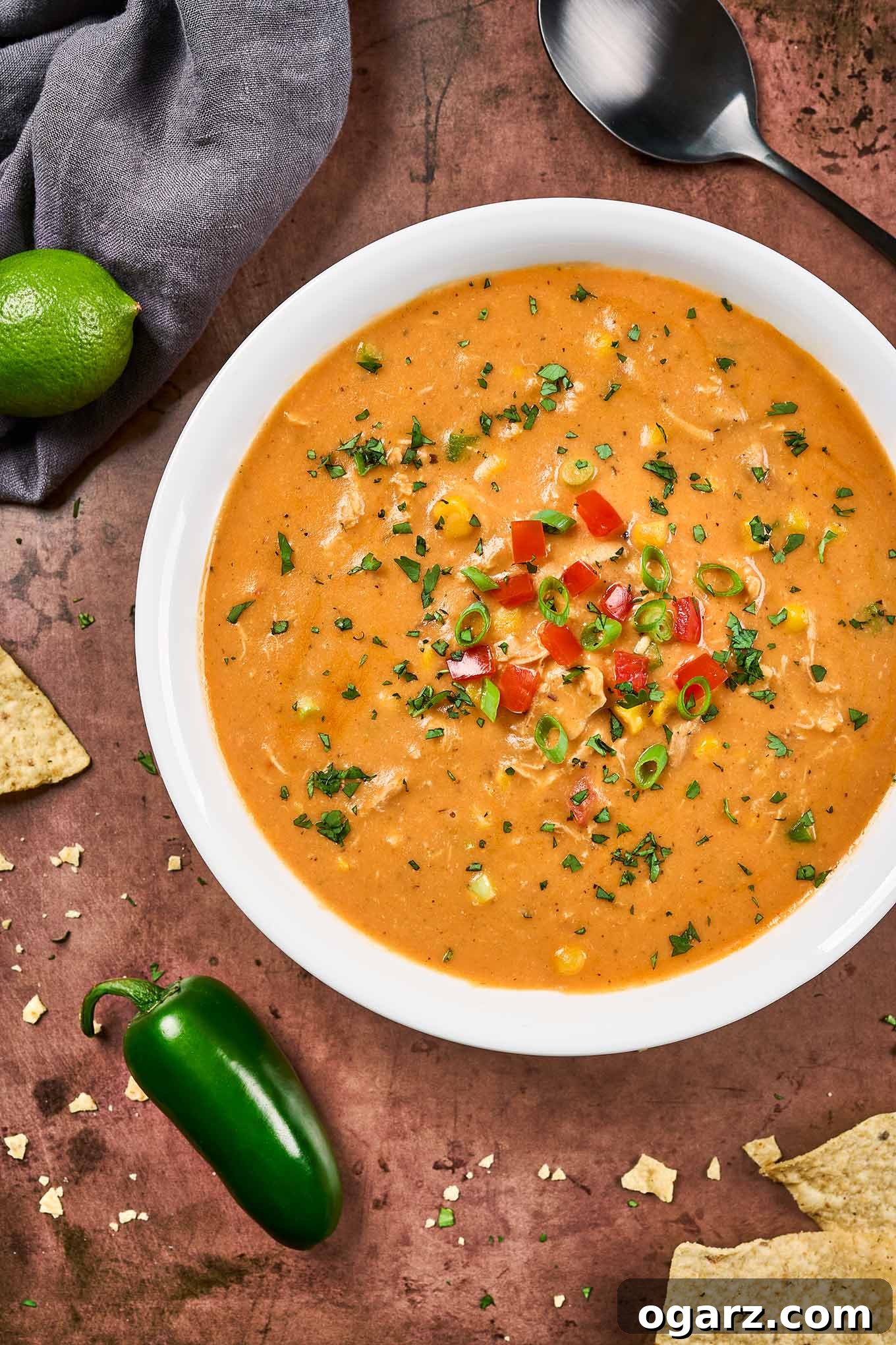 Velvety Chicken Tortilla Soup 4 A bowl of tortilla soup, surrounded by a fresh jalapeno pepper, tortilla chips, a whole lime, and a black soup spoon, all on a distressed brown background, highlighting the fresh garnishes.