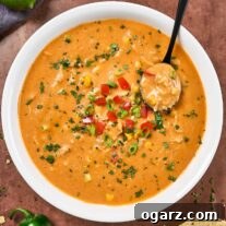 A round white bowl of creamy chicken tortilla soup with a black spoon scooping up a spoonful, a linen napkin, a lime, and tortilla chips in the background.