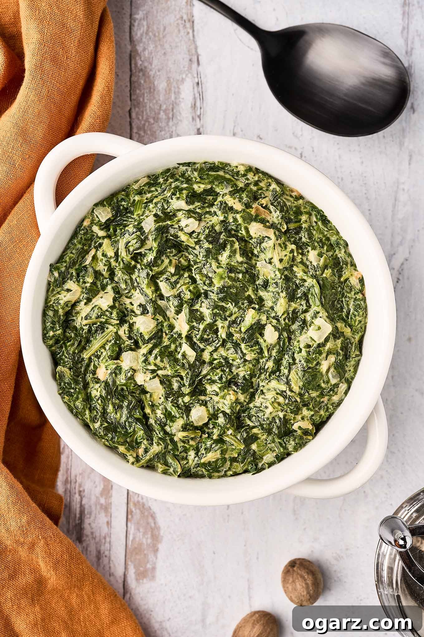 Velvety Creamed Spinach 2 A round, white serving bowl filled with creamed spinach, surrounded by a linen napkin, a large black serving spoon, whole nutmeg, and a nutmeg grinder.