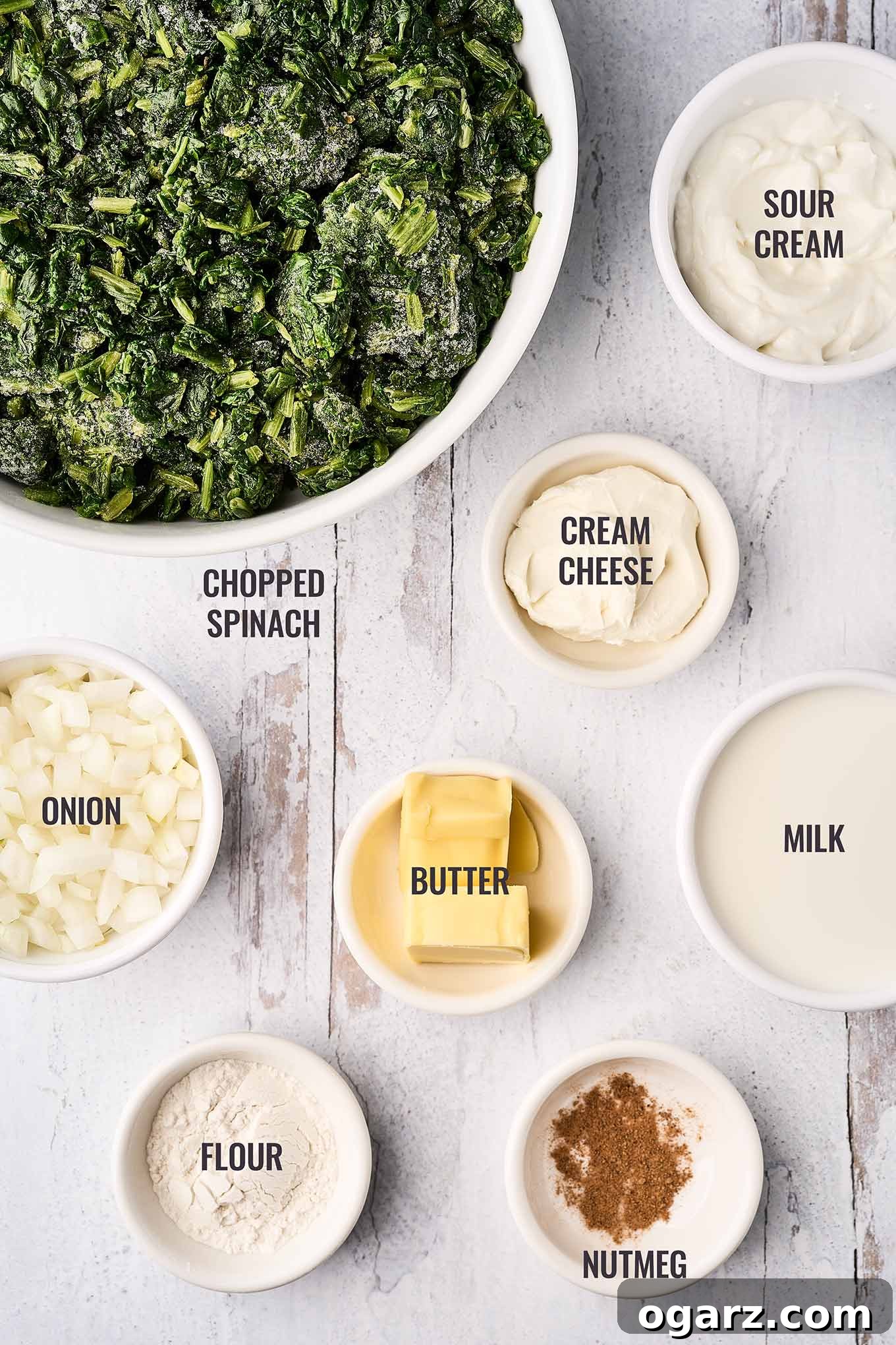 Velvety Creamed Spinach 3 A labeled, overhead view of the ingredients needed to make creamed spinach including chopped spinach, onion, sour cream, cream cheese, and nutmeg.