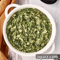 A round, white serving bowl filled with creamed spinach, surrounded by a linen napkin, a large black serving spoon, whole nutmeg, and a nutmeg grinder.
