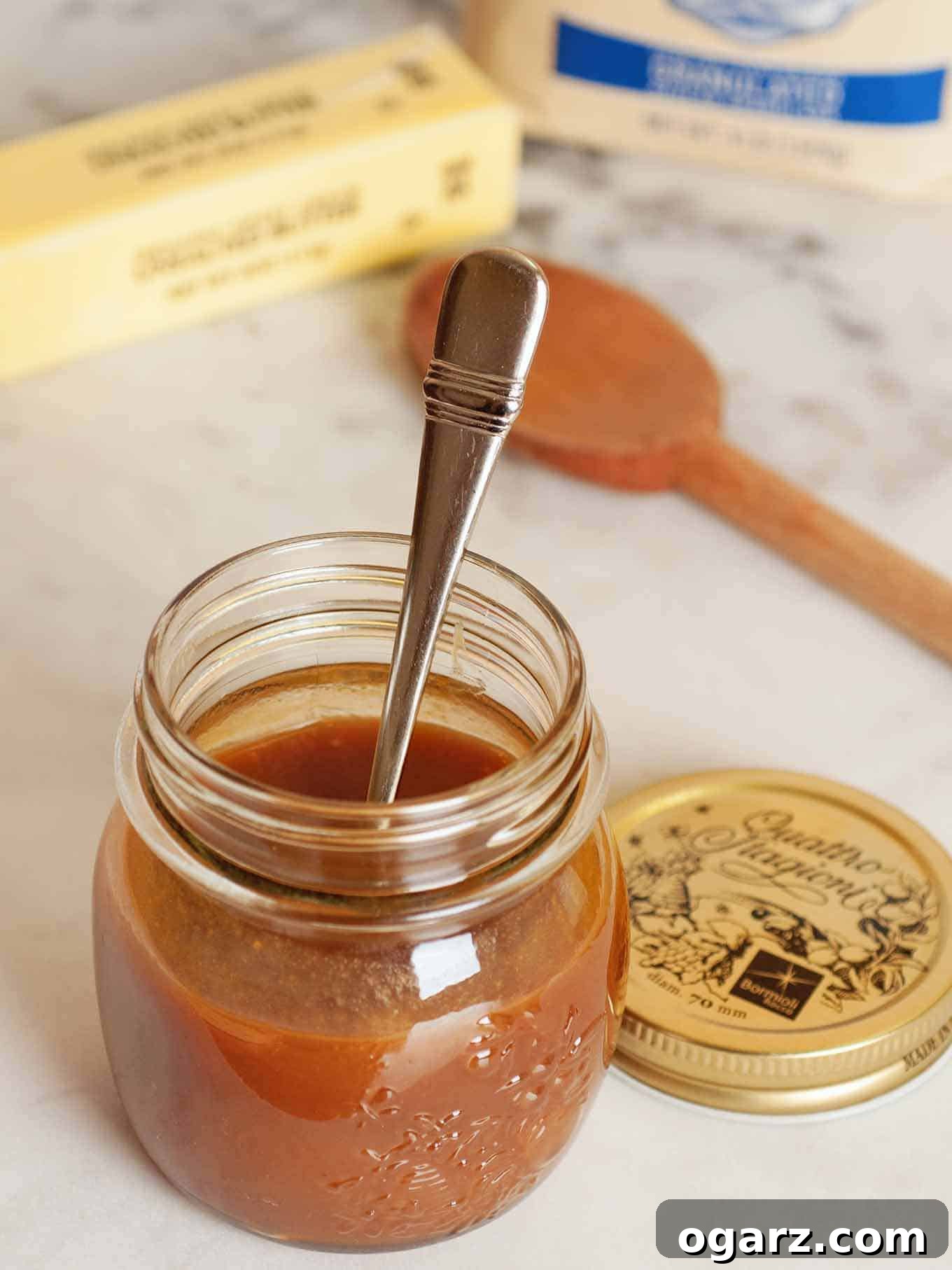 A jar of rich, homemade caramel sauce with a spoon, surrounded by ingredients like butter, sugar, and heavy cream on a marble countertop.