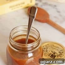 A jar of homemade caramel sauce with a spoon inside, on a marble countertop with a stick of butter, bag of sugar, and wooden spoon in the background.