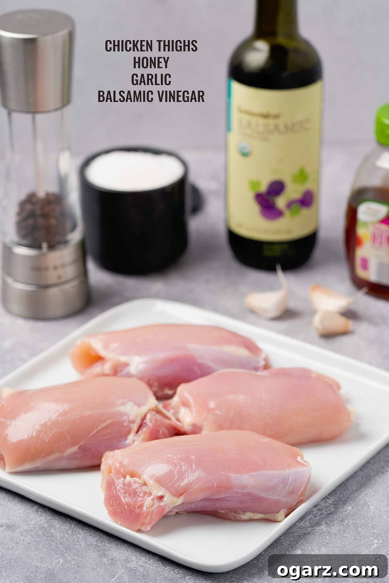 Golden Roasted Boneless Chicken Thighs 3 A collection of key ingredients for roasted chicken thighs, including boneless, skinless chicken thighs, balsamic vinegar, golden honey, fresh garlic cloves, and a seasoning of salt and pepper.