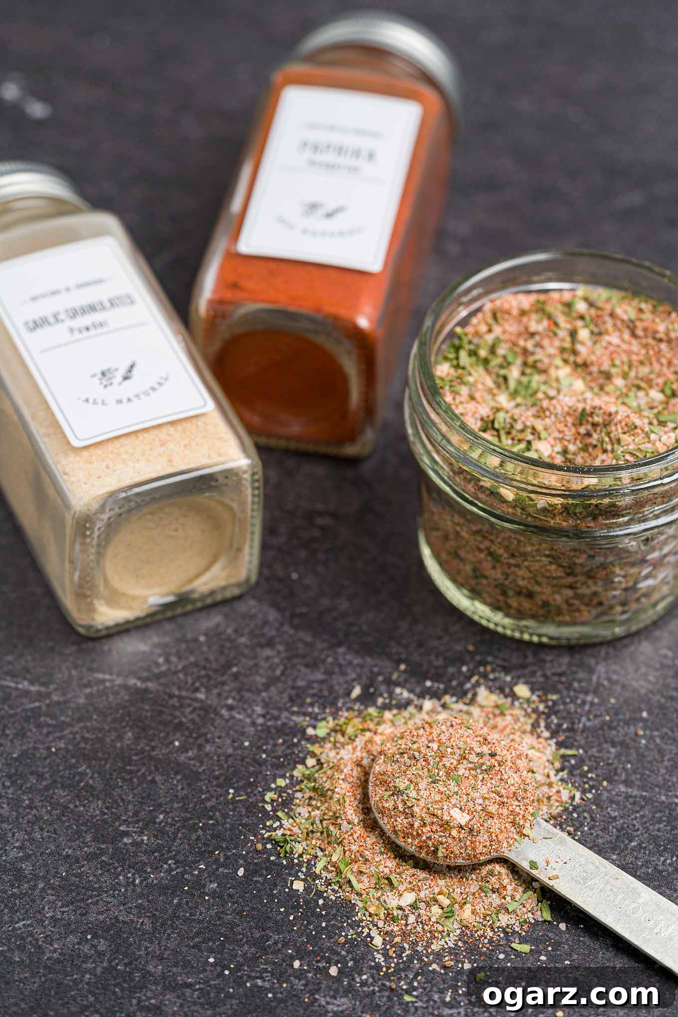 A small jar filled with homemade all purpose seasoning, a measuring spoon in front of it, two spice jars of ingredients in the background.
