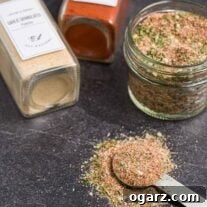 A small jar filled with homemade all purpose seasoning, a measuring spoon in front of it, two spice jars of ingredients in the background.
