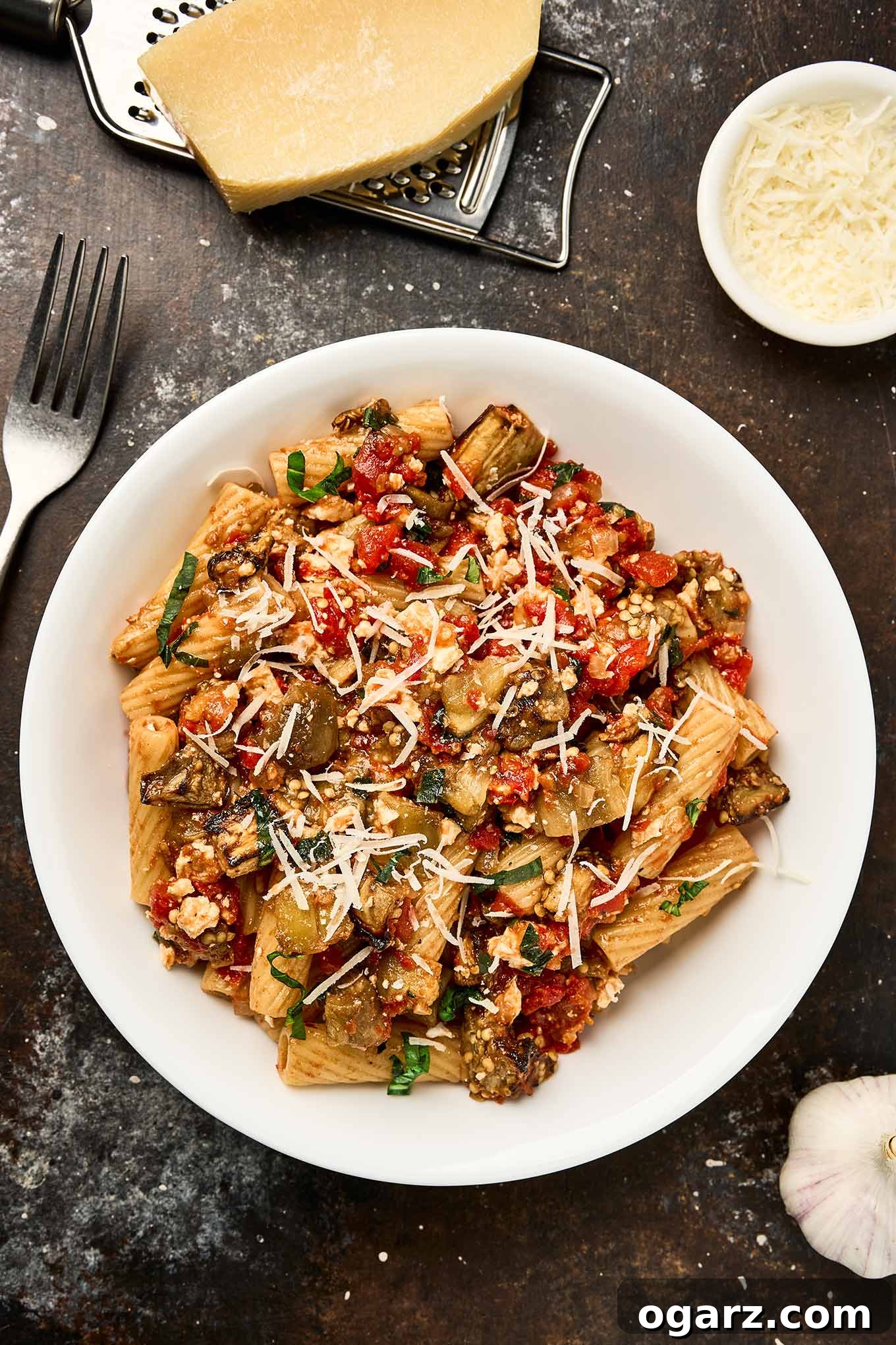Pasta alla Norma A Sicilian Roasted Eggplant Classic 2 A white bowl filled with Pasta alla Norma - pasta tossed with roasted eggplant, tomato sauce, ricotta salata, and basil, topped with shredded cheese. A wedge of cheese, a grater, a fork, and a bowl of grated cheese are arranged nearby.
