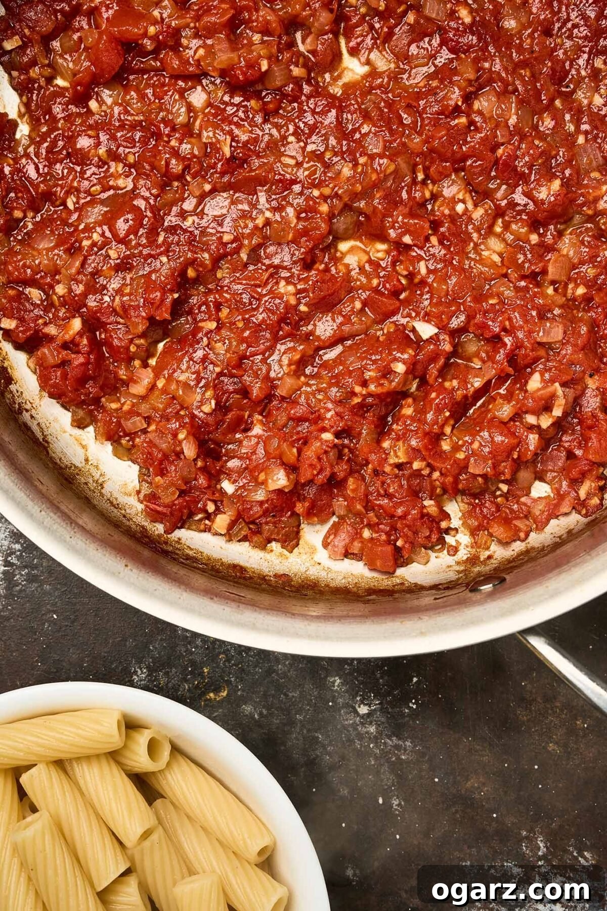 Pasta alla Norma A Sicilian Roasted Eggplant Classic 5 A skillet filled with tomato sauce made with diced tomatoes, garlic, and onions. Below the pan, a bowl of al dente cooked rigatoni pasta is ready to be added to the sauce.