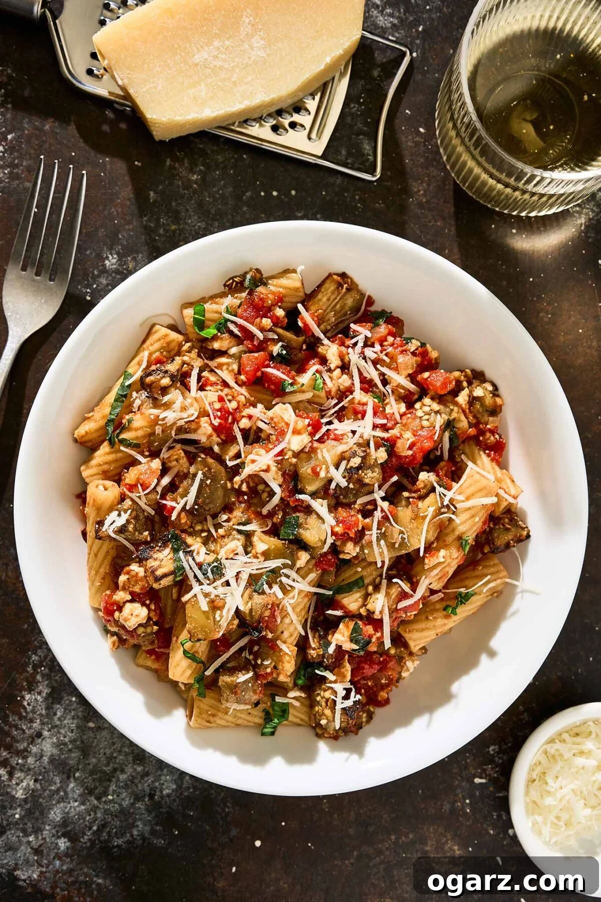 Pasta alla Norma A Sicilian Roasted Eggplant Classic 7 A bowl of rigatoni with roasted eggplant, tomato sauce, ricotta salata, and fresh basil topped with grated Parmesan cheese and surrounded by a wedge of cheese on a grater, a fork, a glass of white wine, and a small bowl of extra grated cheese.