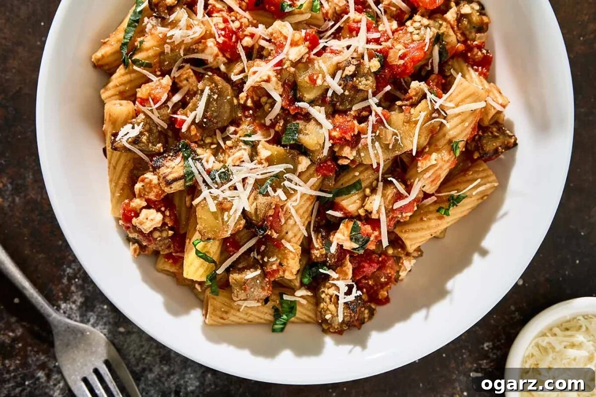 Pasta alla Norma A Sicilian Roasted Eggplant Classic 8 A close-up of Pasta alla Norma in a white bowl, featuring rigatoni tossed with roasted eggplant, chunky tomato sauce, fresh basil, and ricotta salata. A fork and small dish of grated cheese sit nearby on a dark surface.