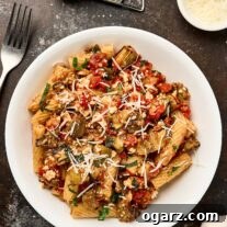 A white bowl filled with Pasta alla Norma - pasta tossed with roasted eggplant, tomato sauce, ricotta salata, and basil, topped with shredded cheese. A wedge of cheese, a grater, a fork, and a bowl of grated cheese are arranged nearby.