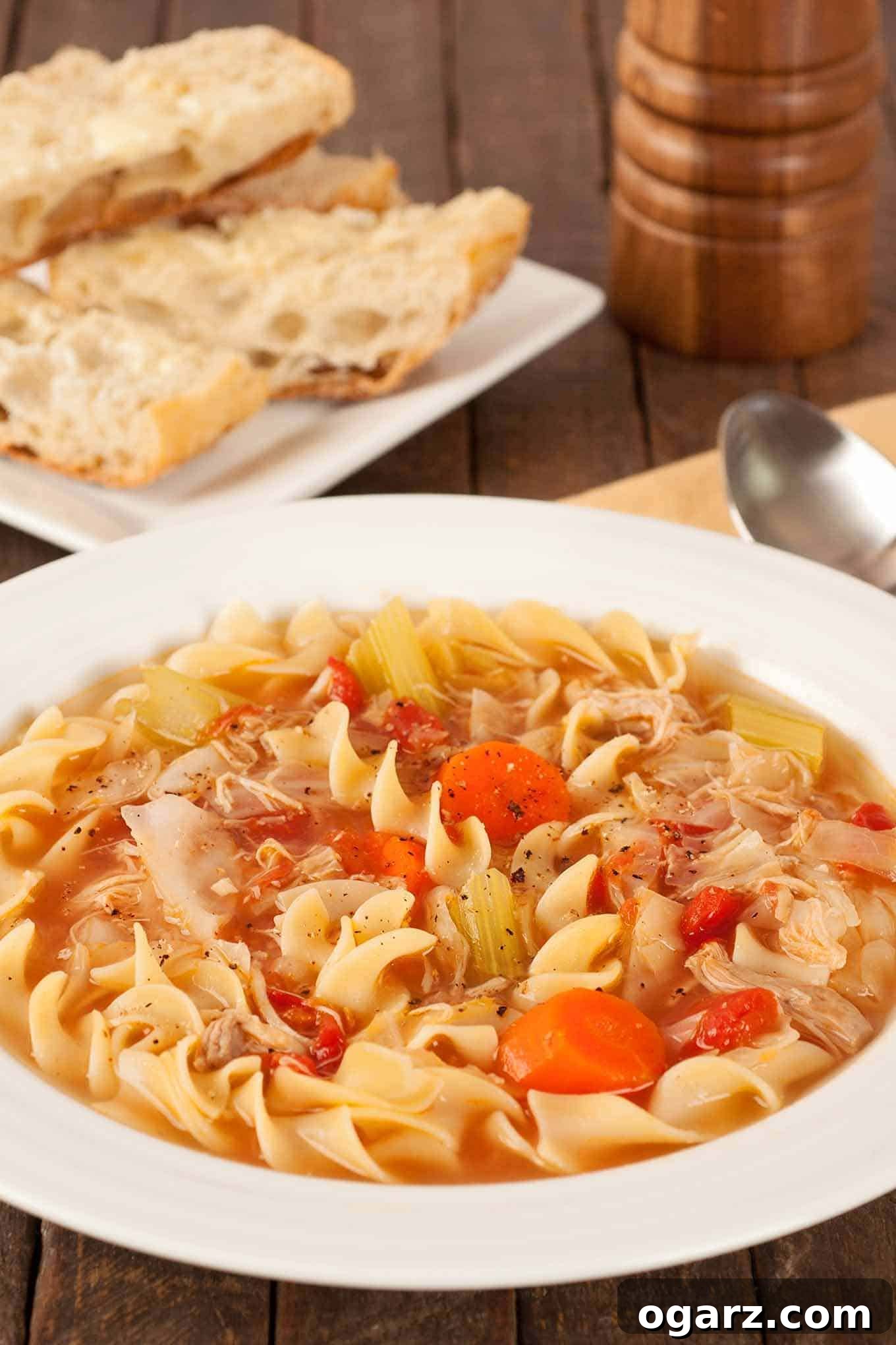 Soulful Chicken Noodle Delight 2 A bowl of chicken soup with curly noodles, chicken, carrots, celery, tomatoes, and cabbage, surrounded by a napkin, spoon, peppermill, and plate with crusty bread.