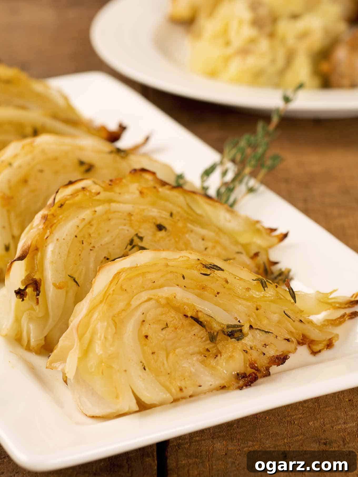 Perfectly Roasted Garlic Herb Cabbage 2 Wedges of golden-brown garlic and herb roasted cabbage on a serving plate, garnished with fresh thyme, with a dinner plate in the background.