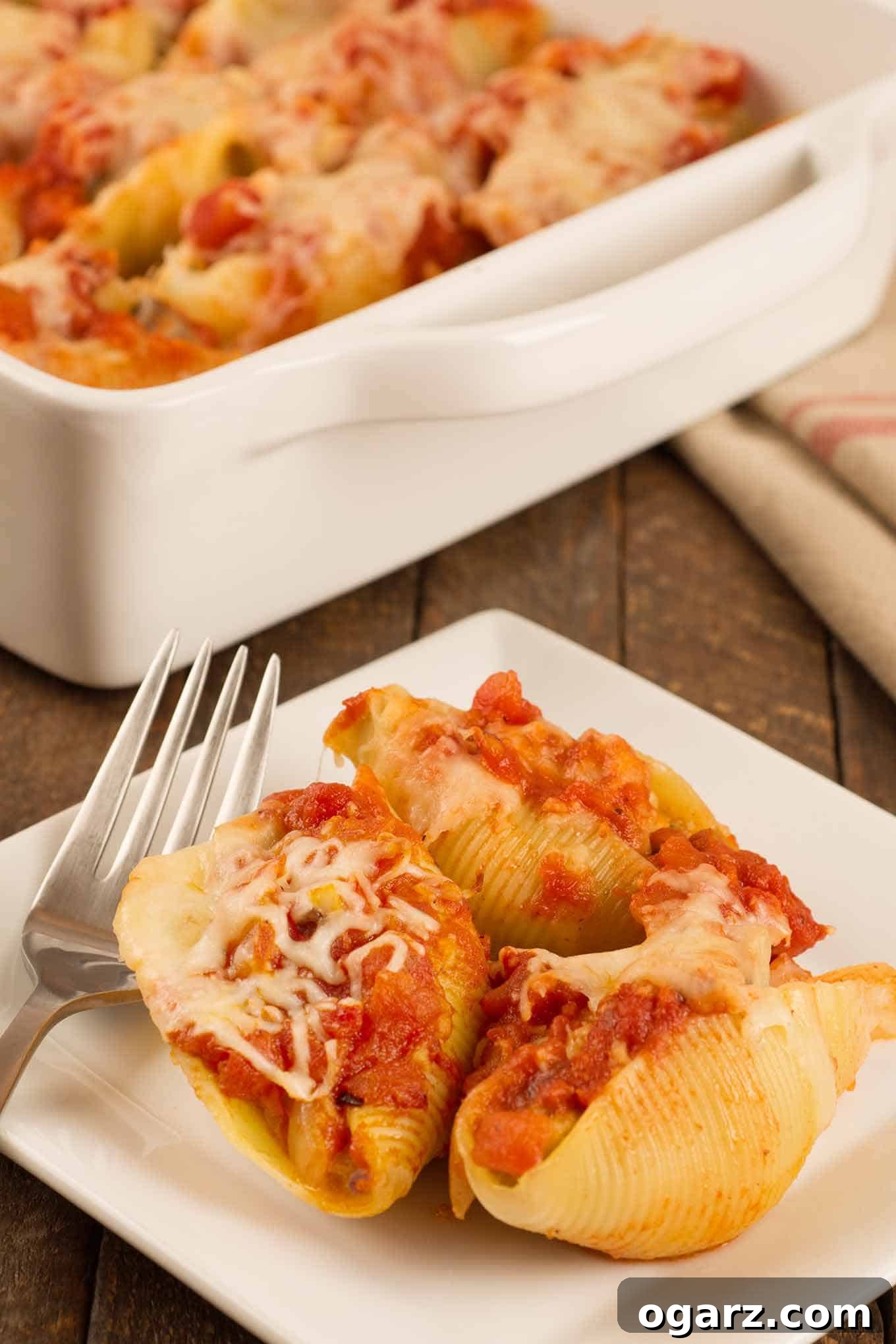 Hearty Sausage Stuffed Shells 2 Sausage stuffed pasta shells topped with marinara and melted cheese, served on a white plate with a fork, the casserole with more shells in the background.