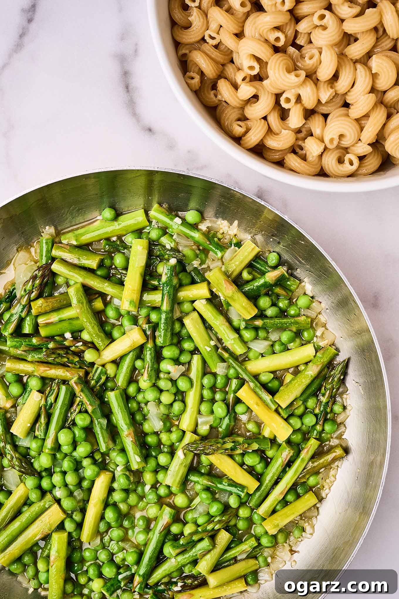 A frying pan filled with sautéed garlic, onion, asparagus, and sweet peas with a bowl of uncooked cavatappi pasta behind it.