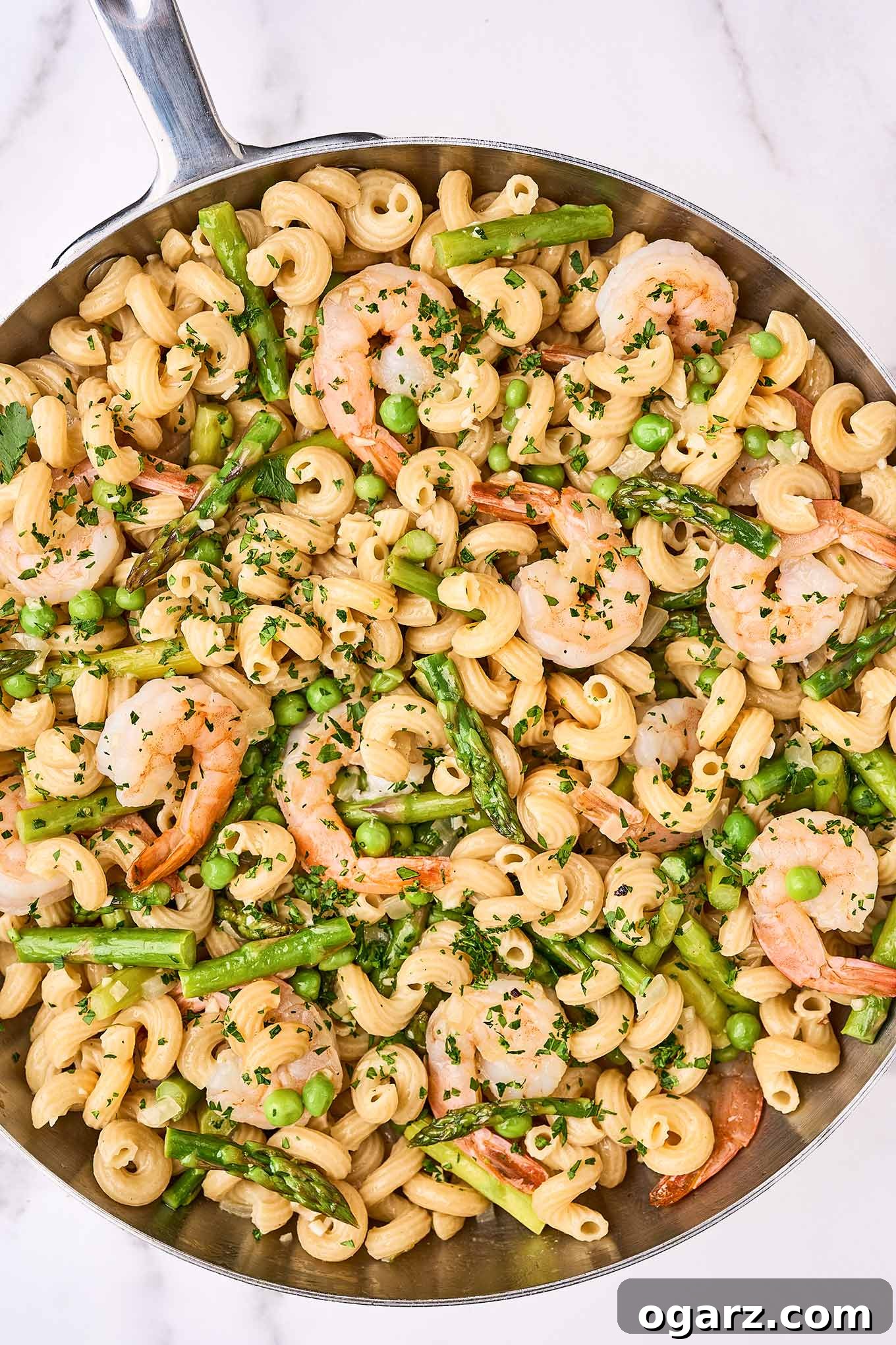 A stainless sauté pan filled with corkscrew pasta combined with sautéed shrimp, asparagus, peas, and parsley, in a light white wine sauce.