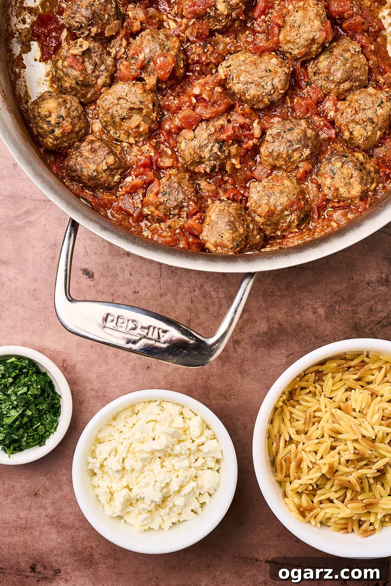 Soutzoukakia: Flavorful Greek Meatballs in Rich Tomato Sauce 12 A sauté pan filled with Greek meatballs in tomato sauce, surrounded by toasted orzo, crumbled feta cheese, and chopped parsley, ready to be plated and served.