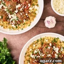 An overhead view of two bowls of Greek meatballs in tomato sauce served over orzo and topped with feta cheese and fresh parsley. A bunch of parsley, bulb of garlic, and bowl of extra feta surround them.