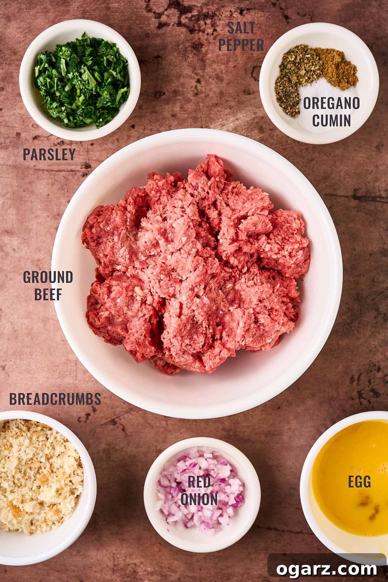 Soutzoukakia: Flavorful Greek Meatballs in Rich Tomato Sauce 3 A labeled, overhead view of the ingredients for Greek meatballs; ground beef, breadcrumbs, egg, red onion, parsley, salt, pepper, oregano, and ground cumin.