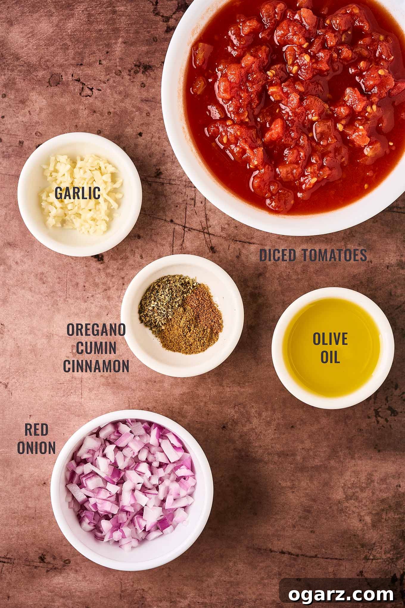 Soutzoukakia: Flavorful Greek Meatballs in Rich Tomato Sauce 4 A labeled, overhead view of the ingredients needed for the tomato sauce for Greek meatballs; diced tomatoes, garlic, olive oil, red onion, dried oregano, ground cumin, and cinnamon.