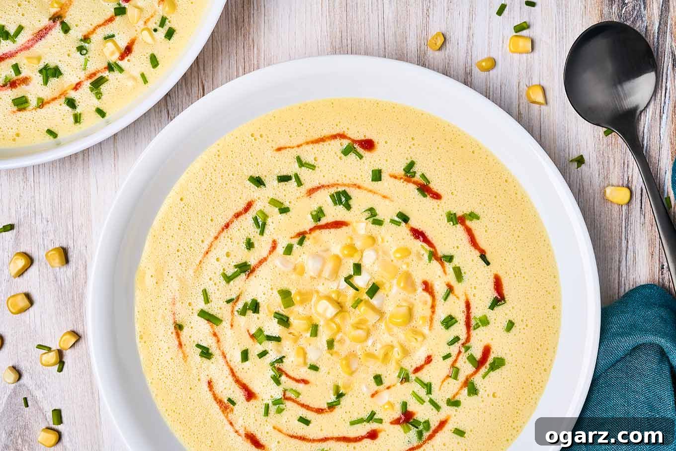 Velvety Sweet Corn Bisque 4 A single elegant serving bowl of golden corn bisque, artfully garnished with a spiral drizzle of red sriracha sauce, a scattering of bright green chopped chives, and a few fresh corn kernels, all set on a light wooden background.