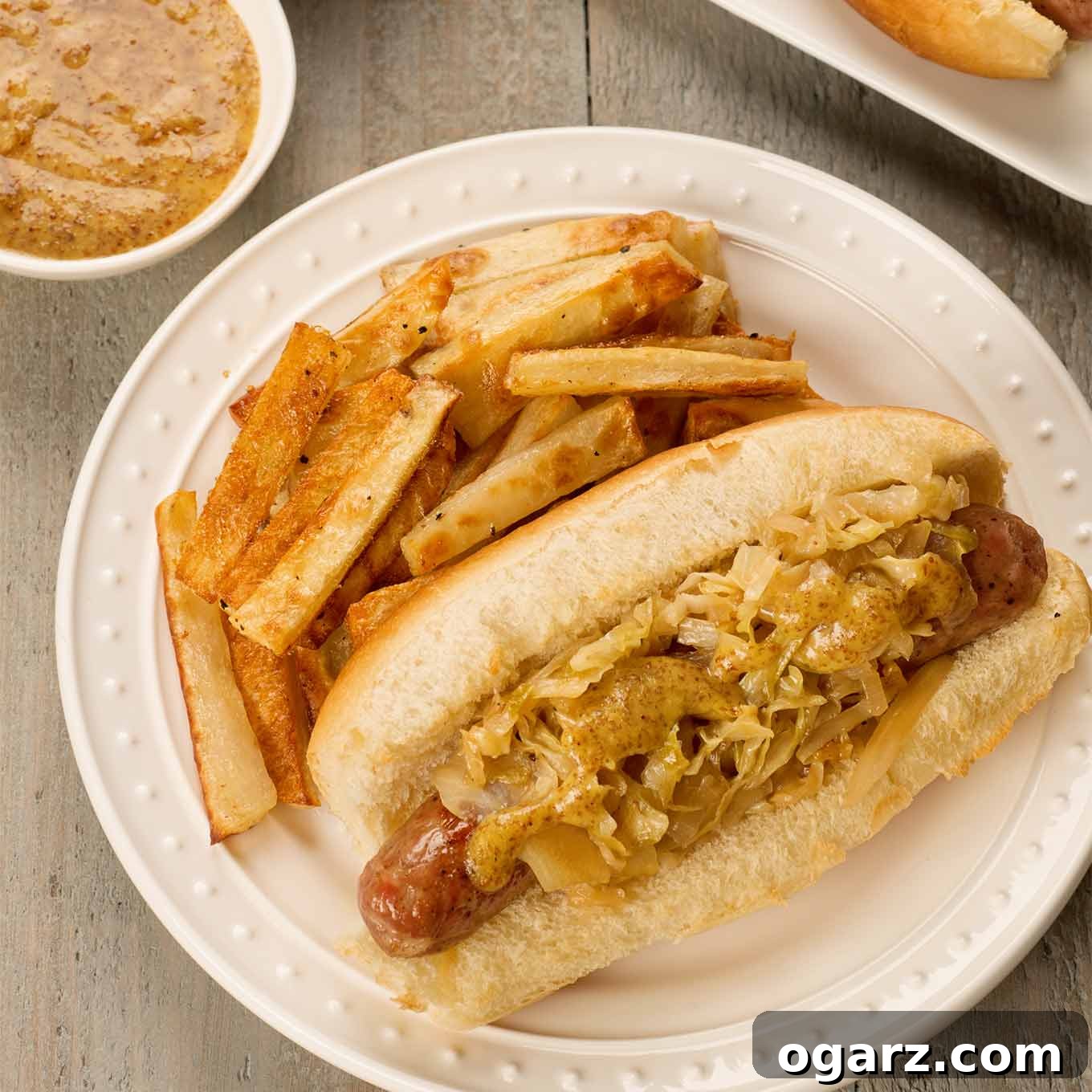 An inviting overhead view of a plated grilled bratwurst on a toasted roll, generously adorned with tender sautéed cabbage and sweet onions, elegantly drizzled with homemade honey mustard. A vibrant side of crispy French fries completes this mouthwatering presentation.