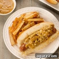 An overhead view of a plate with grilled bratwurst on a roll with sautéed cabbage and onions, a drizzle of honey mustard, and a side of French fries.