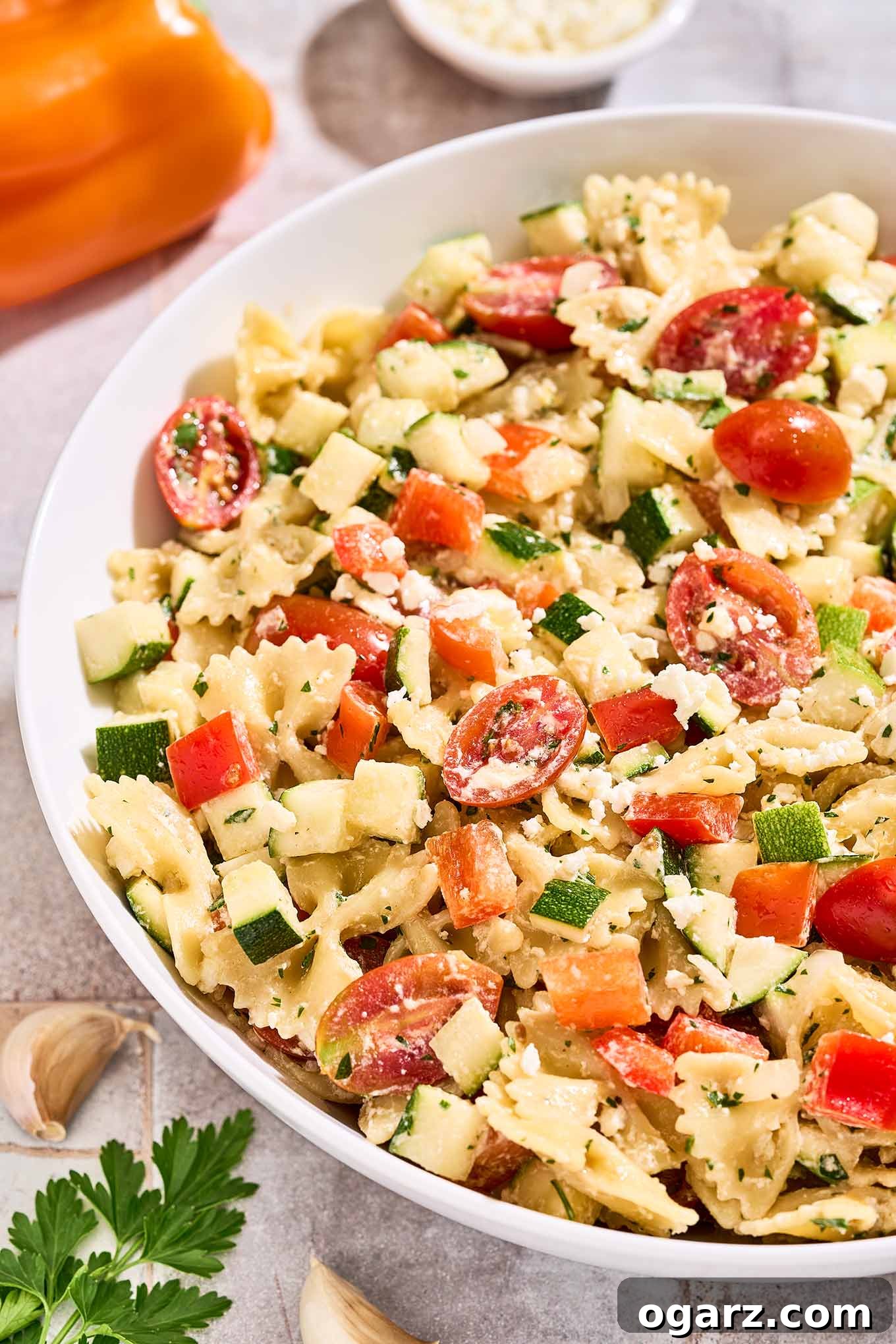 Zesty Lemon Cream Pasta Salad 8 A bowl of creamy pasta salad with lemon dressing surrounded by an orange bell pepper, a dish of crumbled feta, garlic cloves, and a sprig of parsley.