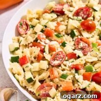 A bowl of creamy pasta salad with lemon dressing surrounded by an orange bell pepper, a dish of crumbled feta, garlic cloves, and a sprig of parsley.